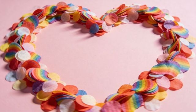 Heart-shaped rainbow confetti wreath on pastel pink backdrop for valentine pride and party