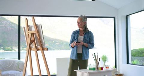 Mature female artist tapping phone in bright coastal studio with flowing color swatches