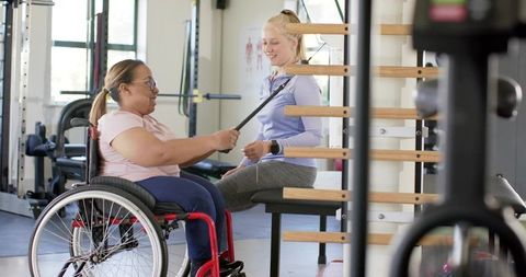 Trainer Assisting Wheelchair User Doing Resistance Band Exercises on Stall Bars
