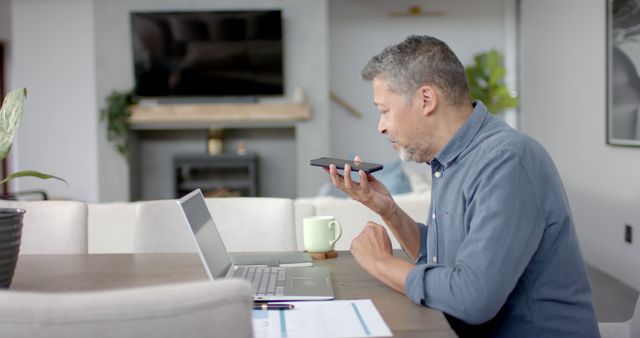 Middle-aged man working from home speaks into smartphone for voice input while sitting in front of laptop. Mug and paperwork on table indicate a productive work environment. This image is great for articles or advertisements focused on remote work, telecommuting, modern technology, and work-life balance.