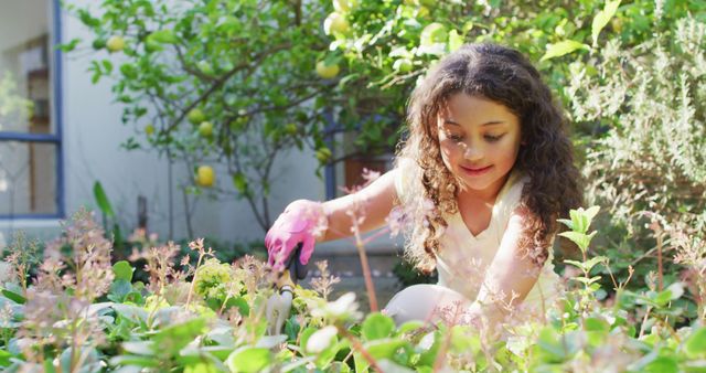 Young girl with curly hair and gardening gloves tending plants in a sunny backyard. Ideal for materials related to outdoor activities, gardening, childhood hobbies, summer leisure, and family home life.