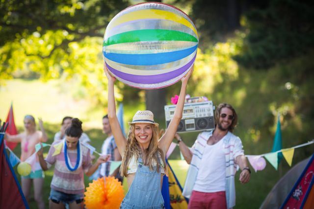 Woman holding a large beach ball above her head at a campsite on a sunny day. She is smiling and surrounded by friends who are enjoying the outdoor setting. The scene is festive with colorful decorations and a boombox, suggesting a lively summer party or festival. Ideal for use in advertisements, travel brochures, summer event promotions, and lifestyle blogs.