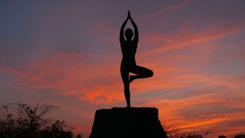 Balancing yoga silhouette practicing tree pose on cliff at vibrant sunset for wellness