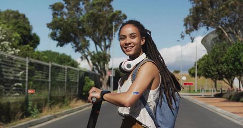 Smiling woman holding electric scooter on campus road wearing backpack and headphones