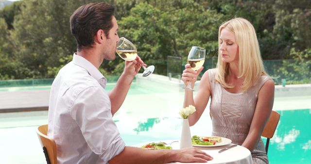 A young Caucasian couple enjoys a romantic outdoor meal by a pool, with copy space. They are toasting with glasses of white wine, enhancing the intimate atmosphere of their dining experience.