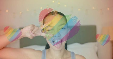 Playful woman making v sign winking and sticking out tongue with rainbow heart overlay