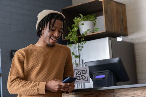 Smiling African American man scanning QR code with phone at cozy brick wood cafe counter