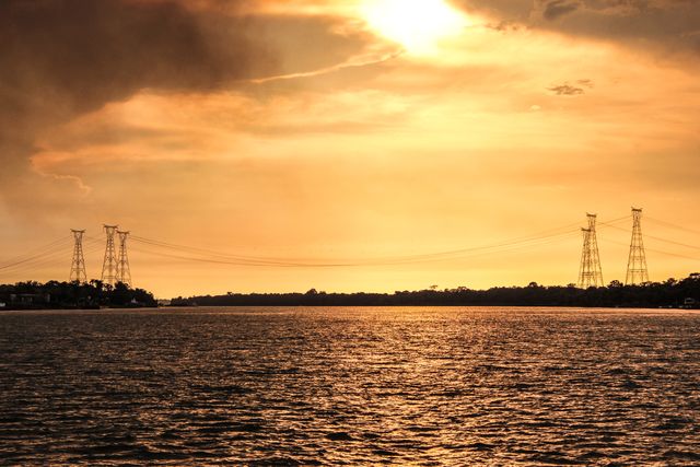 Golden sunset over river with silhouettes of power lines. Sunlight reflects off water creating peaceful and tranquil scene. Perfect for concepts of nature, serenity, tranquility, and electricity. Ideal for environmental themes and backgrounds for websites or presentations highlighting the harmony between nature and industry.