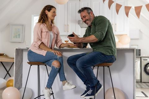 Middle aged couple sharing laughs and phone over wine at decorated kitchen island