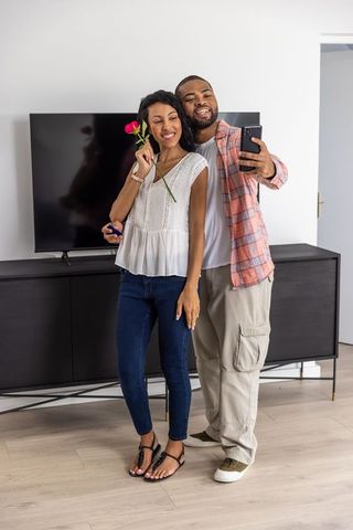 Diverse couple taking selfie while sharing rose and gift box in modern living room