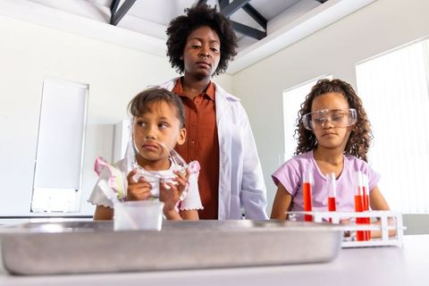 Diverse female science instructor guiding kids through hands-on lab experiment with safety goggles