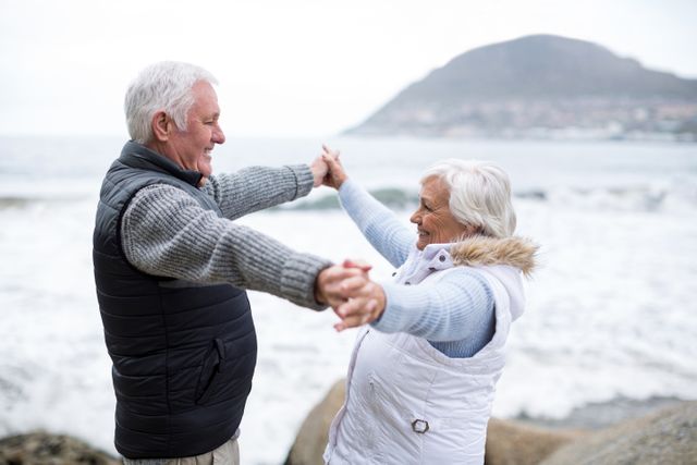 Senior couple smiling and holding hands at the beach with mountains in the background. Perfect for content about aging gracefully, outdoor activities, senior lifestyle, and relationships.