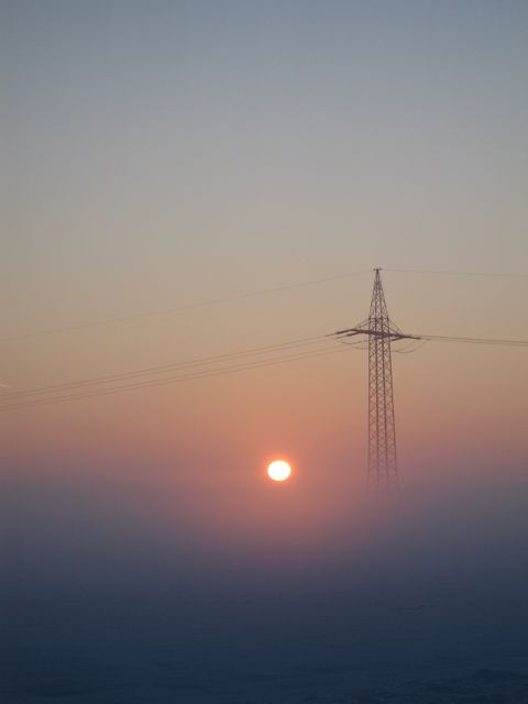 Sunrise casting a warm glow over a misty field with power lines extending into the distance. Ideal for use in energy, renewable resources, or nature-themed projects demonstrating the balance between technology and natural beauty.