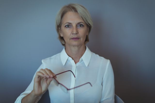 Mature businesswoman holding glasses while sitting against a grey wall. Ideal for corporate presentations, business articles, leadership training materials, and professional profiles.