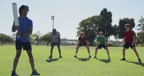 African american batsman taking powerful cricket shot while teens fielding on sunny park