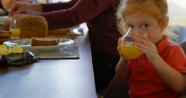 Young girl enjoys drinking orange juice at a family meal, showcasing a warm family moment during breakfast. Ideal for home family life, breakfast scenes, togetherness, and promotional materials for food and beverage products.