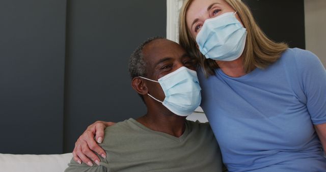 Senior couple sitting on couch at home, smiling and wearing face masks to follow safety measures. Ideal for use in advertisements or articles about health, safety, senior care, wellness, COVID-19 precautions, and diverse relationships.