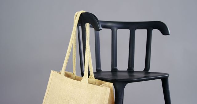Eco-friendly tote bag made of natural jute material hanging on the back of a modern black chair. The image showcases sustainable living and minimalist interior decor, ideal for content promoting reusable bags, environmentally-friendly products, or modern lifestyle concepts.
