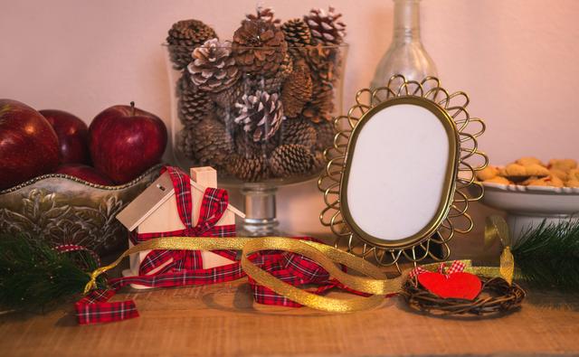Christmas decorations arranged on a wooden table featuring pinecones, red apples, ribbon-wrapped gifts, and an empty gold frame. Ideal for illustrating holiday home decor, festive ambiance, or Christmas preparation themes.