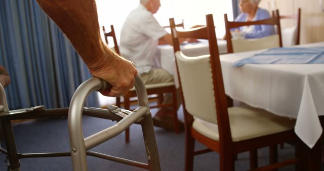 Depicts a senior individual using a walker in a dining room of a care facility. Could be used in healthcare, senior care, and aged care promotional materials or websites to illustrate mobility assistance and senior living environments.