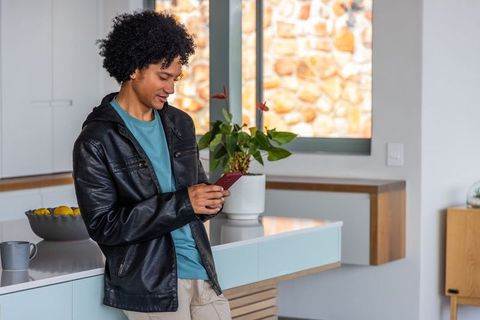 African American man using smartphone in modern sunlit kitchen leaning on island