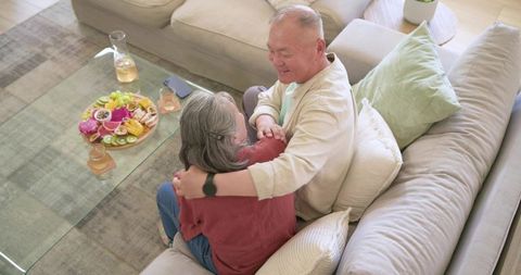 Senior couple embracing on cozy sectional sofa at home with fruit platter and drinks