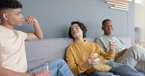 Three young men relaxing on sofa sharing chips and drinks livingroom conversation