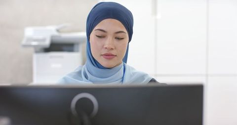Professionally focused muslim woman in navy light-blue hijab working at office desk viewing monitor