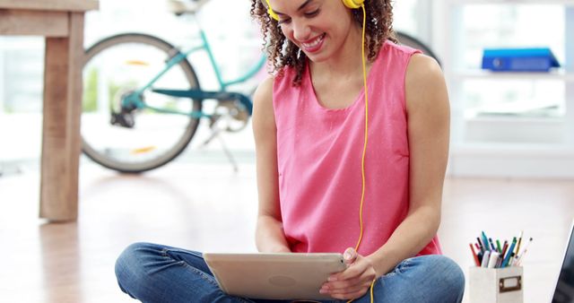 Young woman sitting cross-legged on floor, using a digital tablet and wearing yellow headphones, smiles while browsing or engaging in online activities. Cozy home environment with modern amenities including a bicycle in the background. Ideal for themes like tech-savvy lifestyle, home leisure, remote work, or modern relaxation.