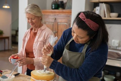 Two women piping frosting on layered cake in cozy kitchen, baking together, sharing joy