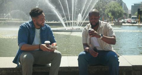 Multiracial friends sharing coffee at urban fountain while checking smartphone outdoors