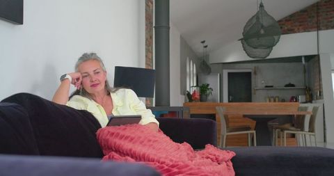 Mature woman relaxing on sofa with tablet in modern open-plan home interior, coral throw