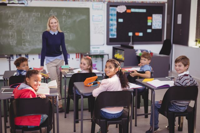 This image shows a smiling teacher standing in front of a diverse group of elementary school students who are sitting at their desks in a classroom. The children are engaged and happy, with books and notebooks on their desks. The chalkboard in the background suggests a traditional classroom setting. This image can be used for educational content, school promotions, teaching materials, and articles about diversity and inclusion in education.