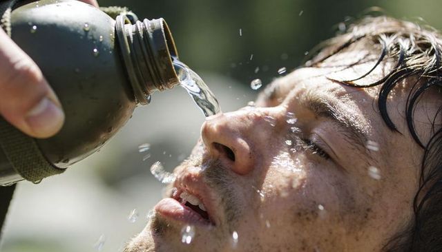 Thirsty hiker drinking from olive-green canteen, tilting face up, water splashing close-up