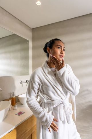 Asian woman applying skincare while wearing white bathrobe in minimalist bathroom vanity