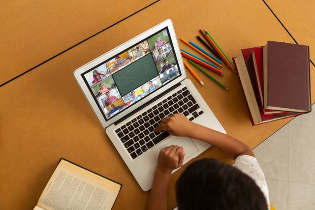 Young student attending virtual class on laptop with multiple participants visible on screen. Surrounding table filled with educational materials, including books and colored pencils. Ideal for illustrating concepts of remote education, home schooling, digital classrooms, and student study environments.