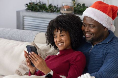 African american couple cuddling on sofa wearing santa hat and sharing cozy holiday smartphone momen