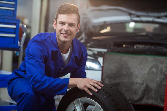 Mechanic in blue uniform smiling while holding a tire in an auto repair garage. Ideal for use in automotive service advertisements, repair shop promotions, and articles on car maintenance and mechanical professions.