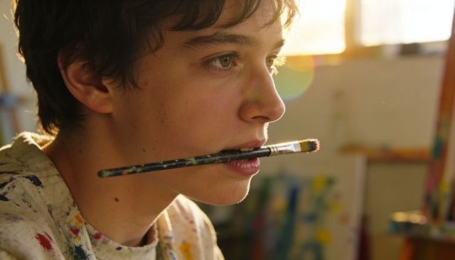 Teen male artist holding paintbrush in mouth gazing toward window light in art studio
