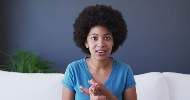 African American woman with afro hair sitting on white couch, looking surprised, indoors at home. Useful for content related to reactions, emotions, casual settings, and relatable everyday scenarios, as well as articles, blogs, and advertisements targeting contemporary living and authenticity.