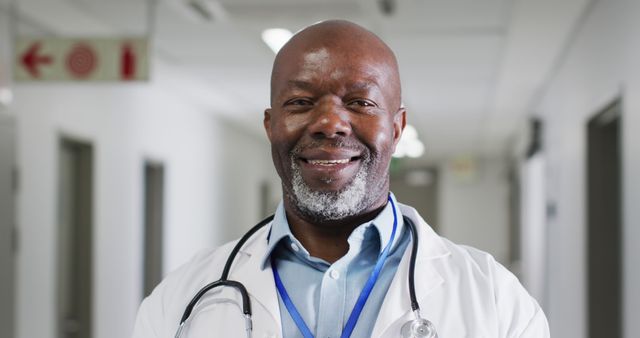 Confident African American doctor smiling in hospital corridor wearing a stethoscope around the neck. Ideal for medical services advertisements, healthcare promotion, patient brochures, medical blog posts, and professional health-related presentations.