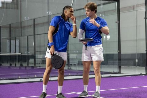 Diverse male doubles team discussing strategy on indoor purple padel court with glass walls