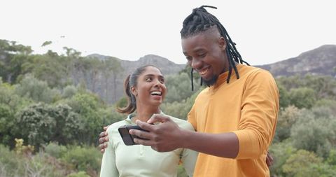 Smiling African American man and Indian woman checking phone on mountainside hike