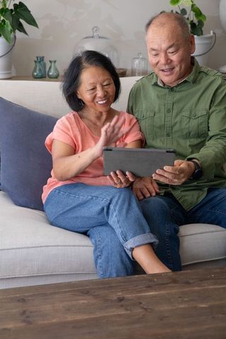 Senior asian couple sharing tablet on cozy sofa, smiling and video calling at home, sunlit living ro