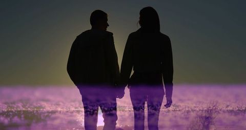 Silhouetted couple holding hands on wet sand at sunset with purple horizon glow