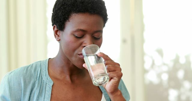 This image depicts a woman taking a drink of water from a glass, highlighting the importance of staying hydrated. Perfect for use in health, wellness, and lifestyle publications or websites promoting the benefits of hydration, natural living, and home-based activities.