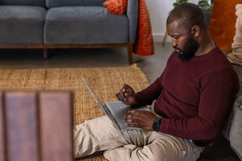 African american man working from home on laptop sitting on living room rug