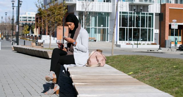 Woman in hijab wearing casual attire sitting on a bench in modern urban park using smartphone. Pink handbag beside her. Ideal for illustrating concepts related to technology, communication, urban lifestyle, cultural diversity, and women empowerment.