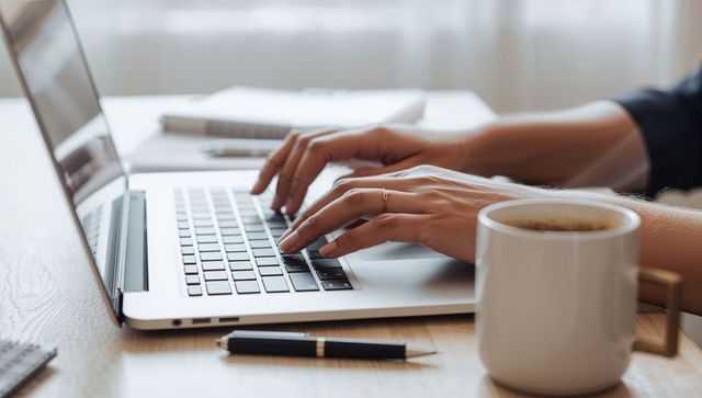 Woman typing on laptop with coffee at minimalist wooden desk, close-up productivity scene
