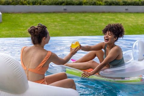 Multicultural friends laughing, enjoying drinks on inflatable floats, sunny backyard pool